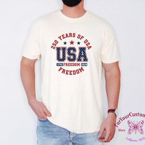 May include: Cream-colored t-shirt with a patriotic design. The design features the text "250 YEARS OF USA" in a circular arrangement, with "USA" prominently displayed in a star-filled font. Additional text includes "1776 FREEDOM 2026 FREEDOM".