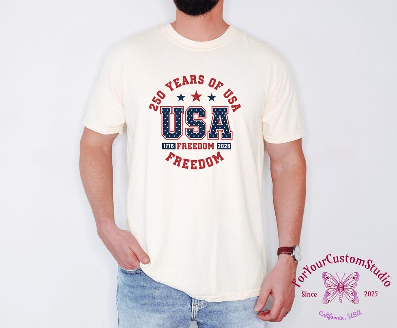 May include: Cream-colored t-shirt with a patriotic design. The design features the text "250 YEARS OF USA" in a circular arrangement, with "USA" prominently displayed in a star-filled font. Additional text includes "1776 FREEDOM 2026 FREEDOM".