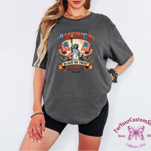May include: Dark gray t-shirt with a patriotic design. The graphic includes the text "250 YEARS OF AMERICA", the Statue of Liberty, American flags, and the words "IN GOD WE TRUST" and "JULY 4th 2026".