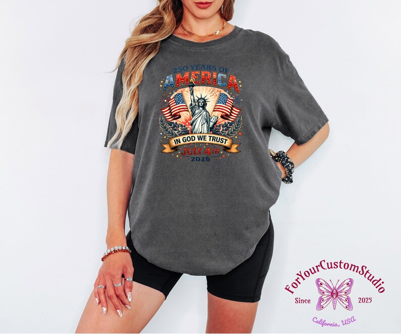 May include: Dark gray t-shirt with a patriotic design. The graphic includes the text "250 YEARS OF AMERICA", the Statue of Liberty, American flags, and the words "IN GOD WE TRUST" and "JULY 4th 2026".