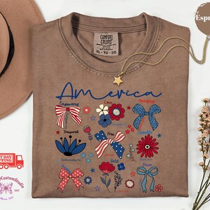 May include: Espresso-colored t-shirt with the word "America" in blue script, adorned with patriotic bow and flower graphics. A gold star pendant necklace rests on the shirt. The shirt is a Comfort Colors XL-TG-EG.