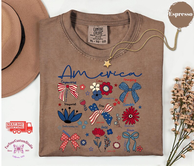 May include: Espresso-colored t-shirt with the word "America" in blue script, adorned with patriotic bow and flower graphics. A gold star pendant necklace rests on the shirt. The shirt is a Comfort Colors XL-TG-EG.