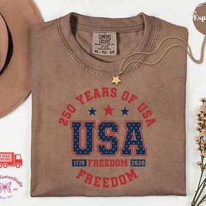 May include: Espresso-colored t-shirt with the text "250 YEARS OF USA" in red and blue. The word "USA" is in a bold, collegiate-style font. The shirt also features the text "1776 FREEDOM 2026 FREEDOM". A gold necklace with a star pendant is draped over the shirt.