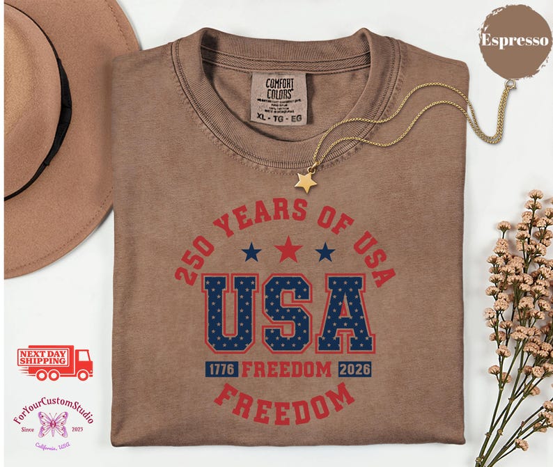 May include: Espresso-colored t-shirt with the text "250 YEARS OF USA" in red and blue. The word "USA" is in a bold, collegiate-style font. The shirt also features the text "1776 FREEDOM 2026 FREEDOM". A gold necklace with a star pendant is draped over the shirt.