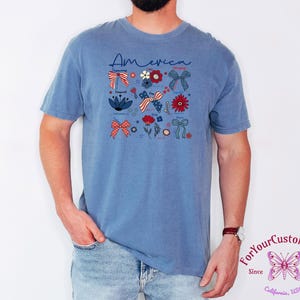 May include: A blue t-shirt with the word "America" in script and patriotic floral and bow graphics. The design includes red, white, and blue elements, with stars and flowers. The shirt is worn by a person.
