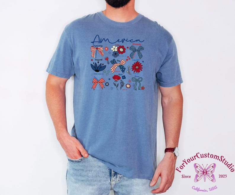 May include: A blue t-shirt with the word "America" in script and patriotic floral and bow graphics. The design includes red, white, and blue elements, with stars and flowers. The shirt is worn by a person.