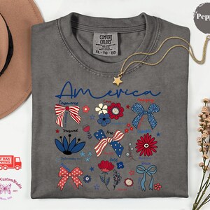 May include: A gray Comfort Colors t-shirt with the word "America" in blue script. The shirt features patriotic bow and floral designs in red, white, and blue. A gold star necklace is draped on the shirt.