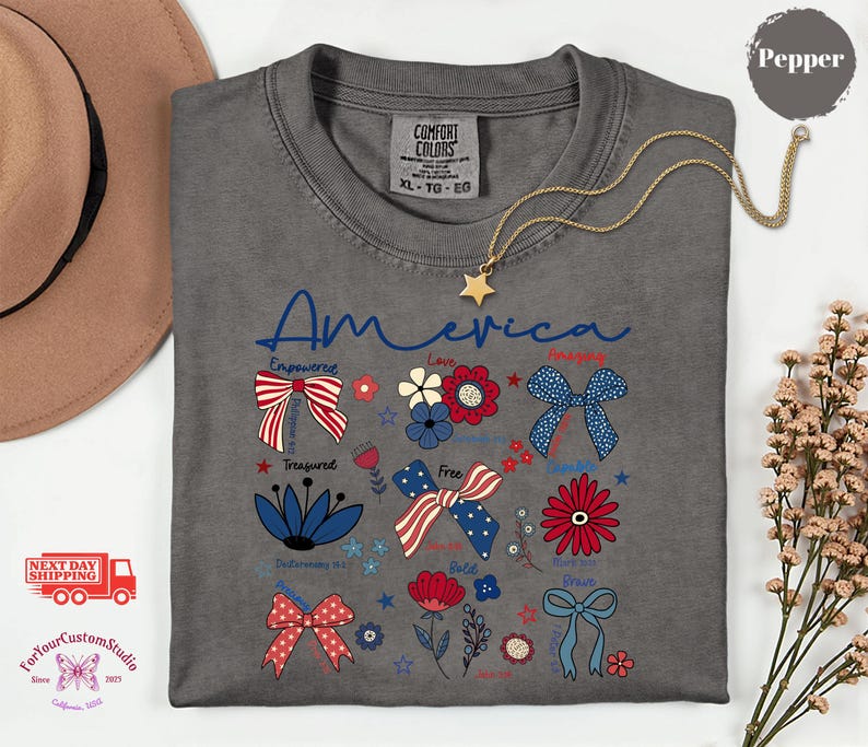 May include: A gray Comfort Colors t-shirt with the word "America" in blue script. The shirt features patriotic bow and floral designs in red, white, and blue. A gold star necklace is draped on the shirt.