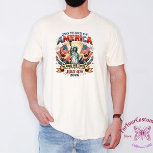May include: Cream-colored t-shirt featuring a patriotic design celebrating 250 years of America. The graphic includes the Statue of Liberty, American flags, and the text "IN GOD WE TRUST" and "JULY 4TH 2026".