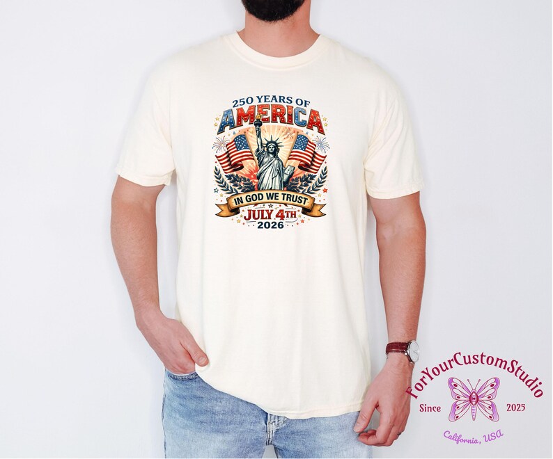 May include: Cream-colored t-shirt featuring a patriotic design celebrating 250 years of America. The graphic includes the Statue of Liberty, American flags, and the text "IN GOD WE TRUST" and "JULY 4TH 2026".