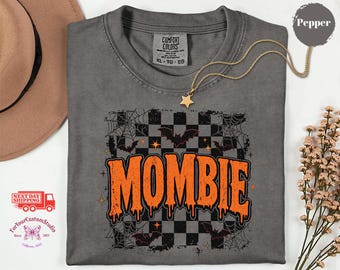 May include: A charcoal gray t-shirt featuring a graphic print with the text MOMBIE in orange, dripping letters. The design includes a black and gray checkered background, spiderwebs, and small bats. The shirt has a Comfort Colors label.