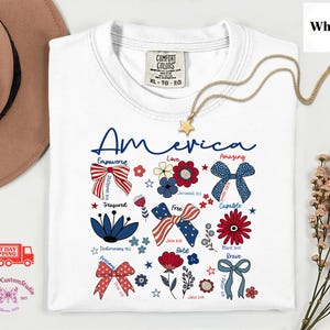 May include: White t-shirt with the word "America" in blue script, featuring patriotic floral and bow designs in red, white, and blue. A gold star pendant necklace is draped across the shirt. The shirt has the Comfort Colors brand tag.