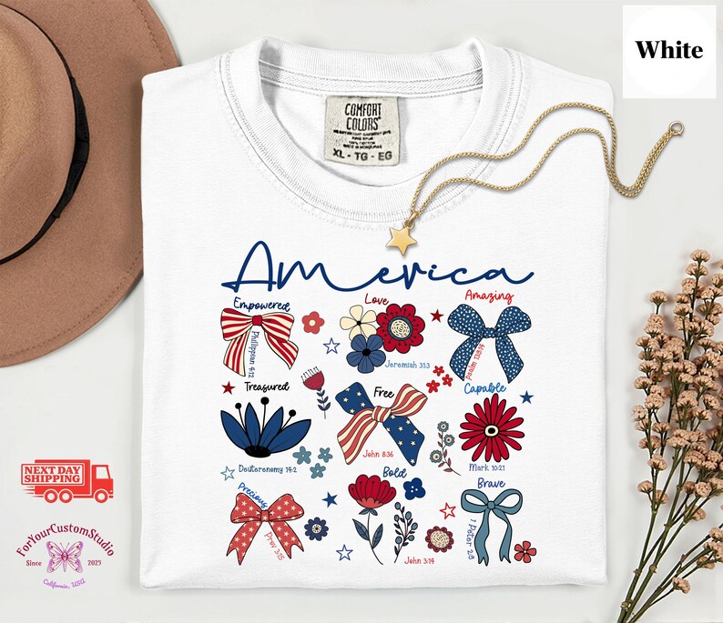May include: White t-shirt with the word "America" in blue script, featuring patriotic floral and bow designs in red, white, and blue. A gold star pendant necklace is draped across the shirt. The shirt has the Comfort Colors brand tag.
