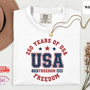 May include: White t-shirt with a patriotic design celebrating 250 years of the USA. The design includes the text "250 Years of USA", "USA" in a blue and white star-patterned font, and the words "1776 Freedom 2026 Freedom" in red.