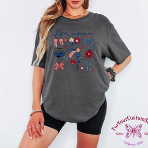 May include: Dark gray t-shirt with the word "America" in blue script and patriotic floral and bow graphics in red, white, and blue. The shirt has a relaxed fit and short sleeves.