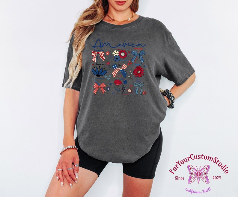 May include: Dark gray t-shirt with the word "America" in blue script and patriotic floral and bow graphics in red, white, and blue. The shirt has a relaxed fit and short sleeves.