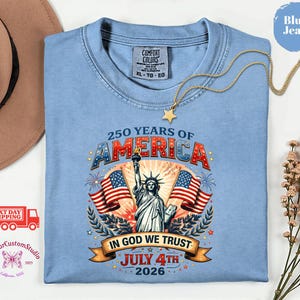 May include: A light blue t-shirt with a graphic celebrating 250 years of America, featuring the Statue of Liberty, American flags, and the text "In God We Trust, July 4th 2026". The shirt is a Comfort Colors XL-TG-EG.