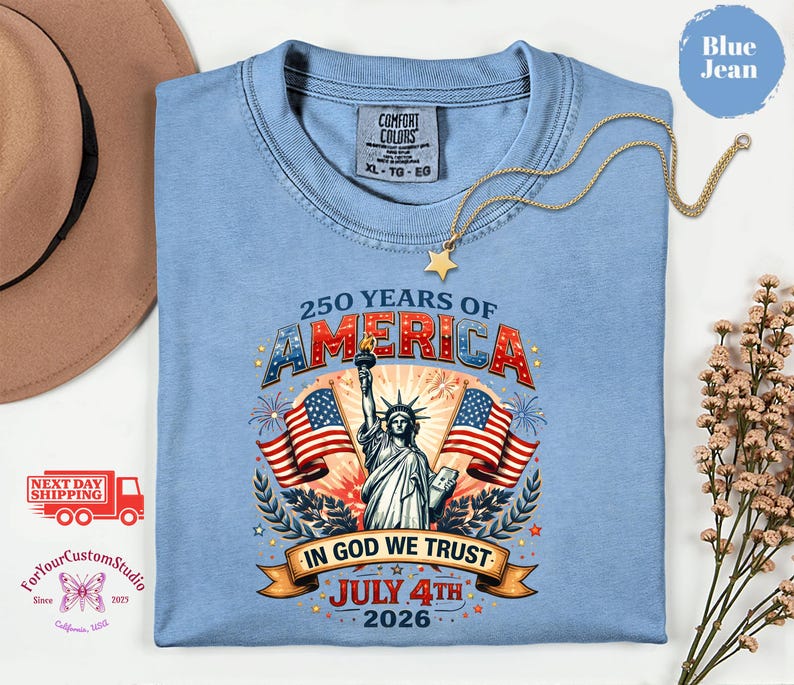 May include: A light blue t-shirt with a graphic celebrating 250 years of America, featuring the Statue of Liberty, American flags, and the text "In God We Trust, July 4th 2026". The shirt is a Comfort Colors XL-TG-EG.