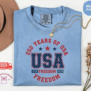 May include: A light blue t-shirt with the text "250 YEARS OF USA" in red and navy blue. The word "USA" is in a bold, patriotic font. The shirt also features the text "1776 FREEDOM 2026 FREEDOM". A gold necklace with a star pendant is draped across the shirt.