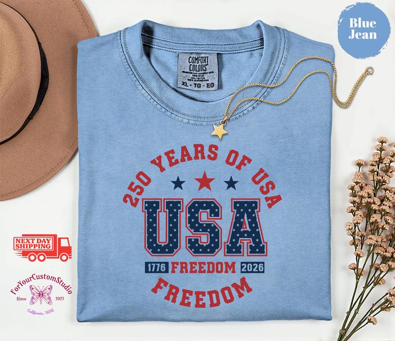 May include: A light blue t-shirt with the text "250 YEARS OF USA" in red and navy blue. The word "USA" is in a bold, patriotic font. The shirt also features the text "1776 FREEDOM 2026 FREEDOM". A gold necklace with a star pendant is draped across the shirt.