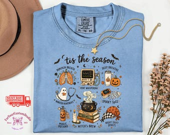 May include: A light blue t-shirt featuring a graphic with the text 'tis the season' and various Halloween-themed medical illustrations including a pumpkin-shaped lung, a heart monitor, a ghost with a stethoscope, and a cauldron on books.