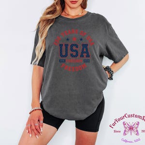 May include: Dark gray t-shirt with a round graphic that reads "250 Years of USA Freedom" in red and blue. The shirt is worn by a person. The graphic includes the years 1776 and 2026.