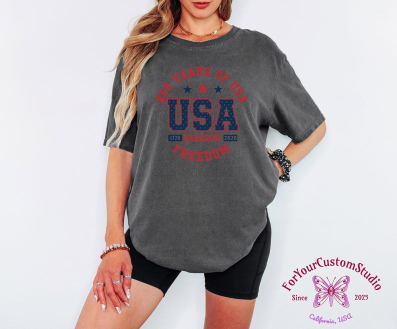 May include: Dark gray t-shirt with a round graphic that reads "250 Years of USA Freedom" in red and blue. The shirt is worn by a person. The graphic includes the years 1776 and 2026.