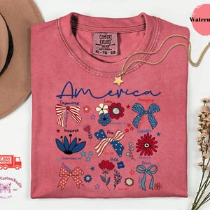 May include: A watermelon-colored t-shirt with the word "America" in blue script. The shirt features patriotic floral and bow designs with words like "Empowered," "Love," and "Free." A gold star necklace is draped across the shirt.