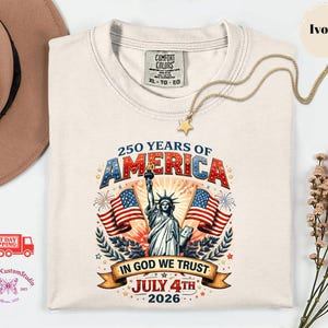 May include: Ivory-colored t-shirt with a graphic celebrating 250 years of America. The design includes the Statue of Liberty, American flags, and the text "In God We Trust" and "July 4th 2026". A gold necklace with a star pendant is draped on the shirt.