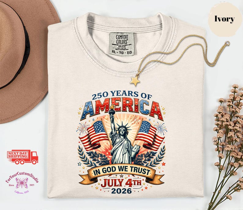 May include: Ivory-colored t-shirt with a graphic celebrating 250 years of America. The design includes the Statue of Liberty, American flags, and the text "In God We Trust" and "July 4th 2026". A gold necklace with a star pendant is draped on the shirt.