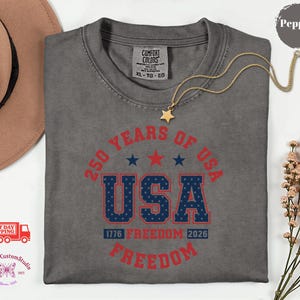 May include: A gray t-shirt with a graphic that reads "250 YEARS OF USA" in red and blue. The shirt also features the text "1776 FREEDOM 2026" and a gold star necklace.
