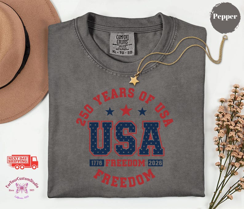 May include: A gray t-shirt with a graphic that reads "250 YEARS OF USA" in red and blue. The shirt also features the text "1776 FREEDOM 2026" and a gold star necklace.