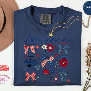 May include: Navy blue t-shirt with the word "America" in script. The shirt features patriotic floral and bow designs in red, white, and blue. A gold star pendant necklace is draped over the shirt.