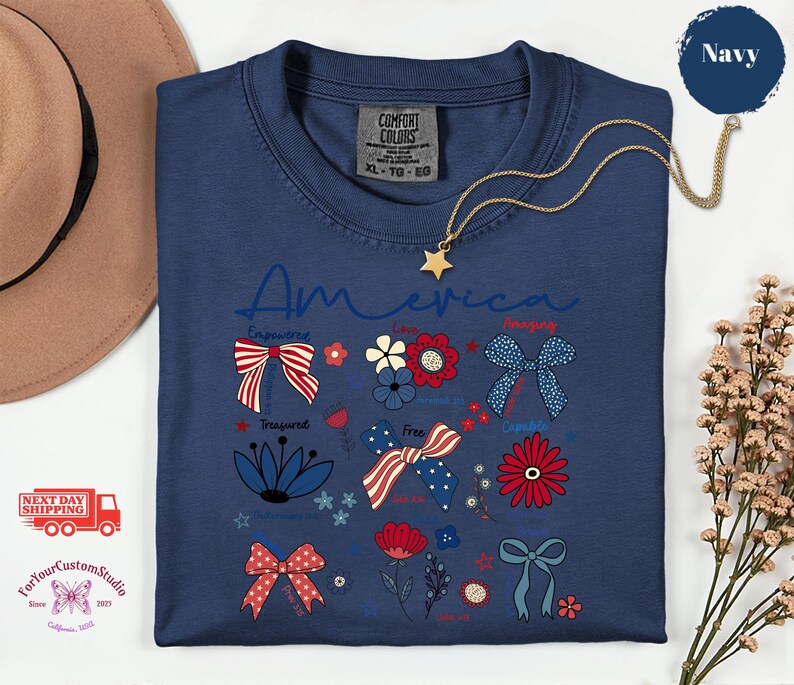 May include: Navy blue t-shirt with the word "America" in script. The shirt features patriotic floral and bow designs in red, white, and blue. A gold star pendant necklace is draped over the shirt.