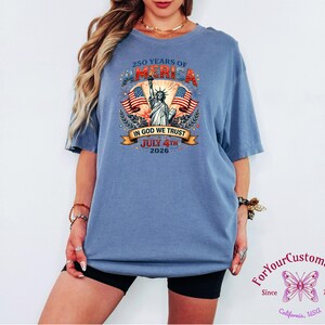 May include: A blue t-shirt with a graphic commemorating 250 years of America, featuring the Statue of Liberty, American flags, and the text "IN GOD WE TRUST" and "JULY 4TH 2026". The shirt also has the text "ForYourCustomStudio" and the year "2025".
