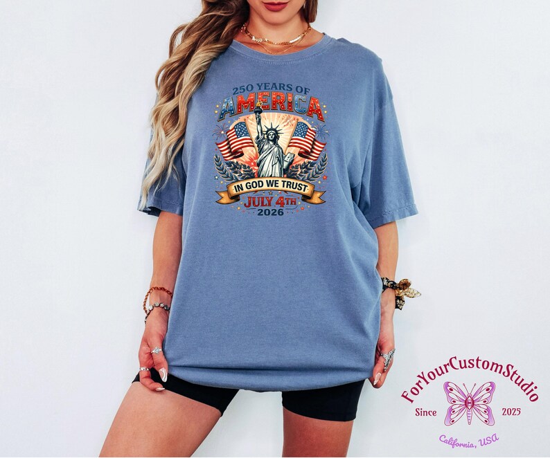 May include: A blue t-shirt with a graphic commemorating 250 years of America, featuring the Statue of Liberty, American flags, and the text "IN GOD WE TRUST" and "JULY 4TH 2026". The shirt also has the text "ForYourCustomStudio" and the year "2025".