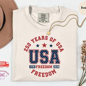 May include: Ivory-colored t-shirt with the text "250 YEARS OF USA" in red and blue. The word "USA" is in a bold, patriotic font. A gold star necklace rests on the shirt. The shirt is a size XL-TG-EG.