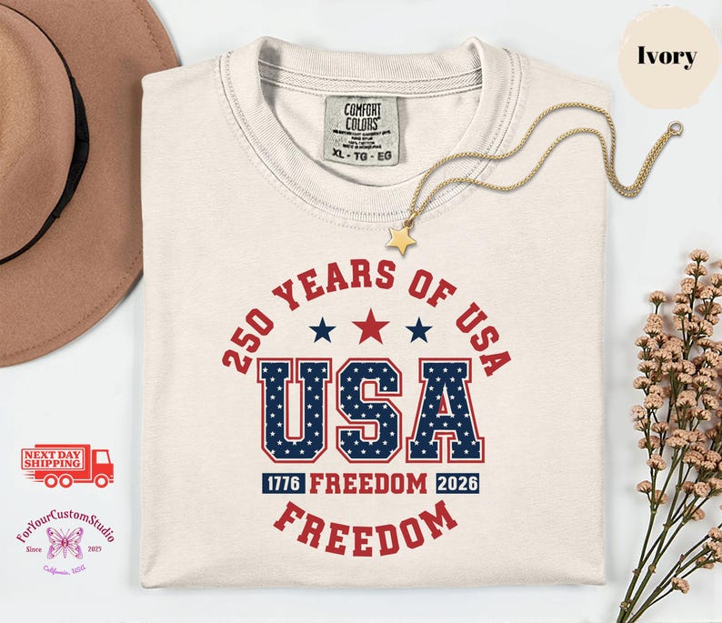 May include: Ivory-colored t-shirt with the text "250 YEARS OF USA" in red and blue. The word "USA" is in a bold, patriotic font. A gold star necklace rests on the shirt. The shirt is a size XL-TG-EG.