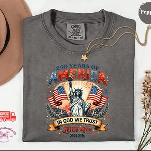 May include: A gray t-shirt with a graphic celebrating 250 years of America. The design features the Statue of Liberty, American flags, and the text "250 Years of America", "In God We Trust", and "July 4th 2026". A gold necklace with a star pendant rests on the shirt.