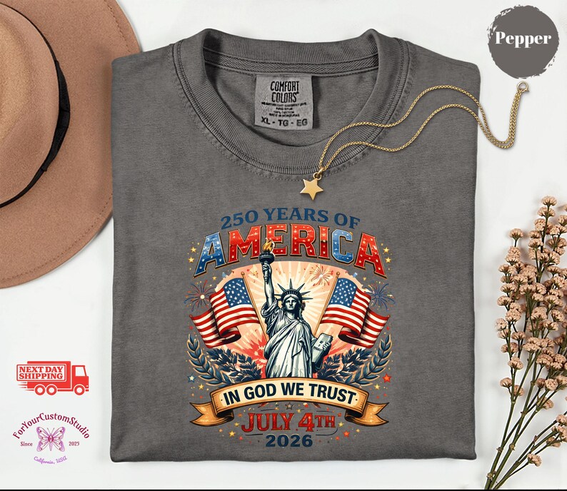 May include: A gray t-shirt with a graphic celebrating 250 years of America. The design features the Statue of Liberty, American flags, and the text "250 Years of America", "In God We Trust", and "July 4th 2026". A gold necklace with a star pendant rests on the shirt.
