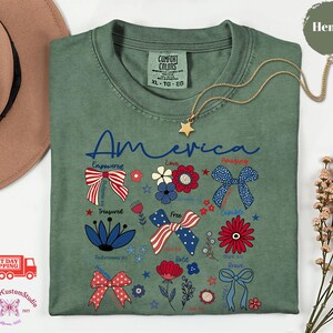 May include: Green t-shirt with the word "America" in blue script, decorated with patriotic bow and flower graphics in red, white, and blue. A gold star pendant necklace rests on the shirt.