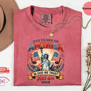 May include: A watermelon-colored t-shirt with a graphic celebrating 250 years of America. The design includes the Statue of Liberty, American flags, and the text "In God We Trust" and "July 4th 2026". A gold star necklace is draped on the shirt.