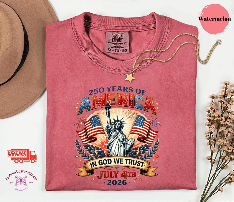 May include: A watermelon-colored t-shirt with a graphic celebrating 250 years of America. The design includes the Statue of Liberty, American flags, and the text "In God We Trust" and "July 4th 2026". A gold star necklace is draped on the shirt.