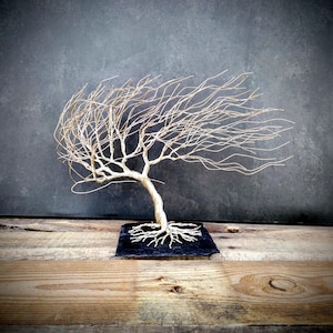 May include: A handcrafted wire tree sculpture with a textured, wind-swept design. The tree features a twisted trunk, delicate branches, and a square black base. The sculpture is displayed on a wooden surface against a dark grey background.