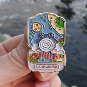 Puede incluir: Un pin de esmalte de color dorado con una rana de dibujos animados sentada en un puente. La rana es azul con la barriga blanca y una concha en espiral en la espalda. El pin representa una pequeña isla con un cuerpo de agua, árboles y una playa. El texto "Seafoam Island" está impreso en la parte inferior del pin.