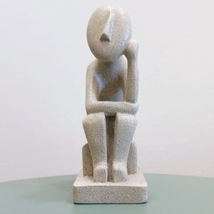Op de afbeelding: Lichtbeige, getextureerde sculptuur van een zittende figuur met het hoofd op de hand. De figuur zit op een rechthoekige basis. De sculptuur heeft een minimalistisch, abstract ontwerp.