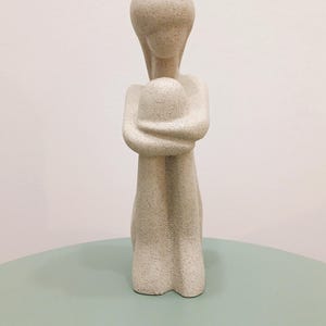 May include: A beige abstract sculpture of a person embracing themselves. The figure is kneeling with arms wrapped around a rounded form. The sculpture has a textured surface and is displayed on a light green surface.