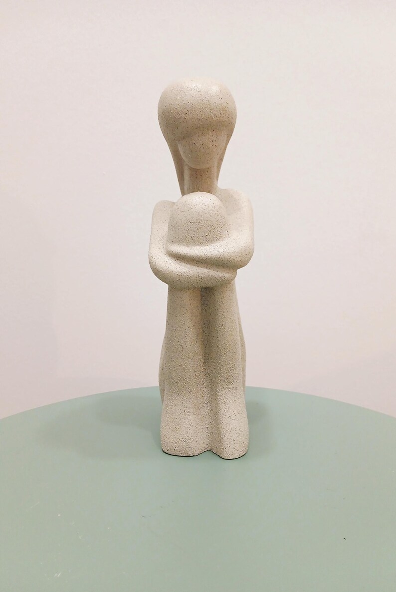 May include: A beige abstract sculpture of a person embracing themselves. The figure is kneeling with arms wrapped around a rounded form. The sculpture has a textured surface and is displayed on a light green surface.