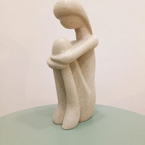 May include: A beige-colored sculpture of a seated figure, knees drawn up to the chest, arms wrapped around the legs. The figure has a smooth, rounded head and body, with a textured surface. The sculpture is on a light green surface.