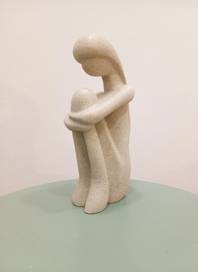 May include: A beige-colored sculpture of a seated figure, knees drawn up to the chest, arms wrapped around the legs. The figure has a smooth, rounded head and body, with a textured surface. The sculpture is on a light green surface.